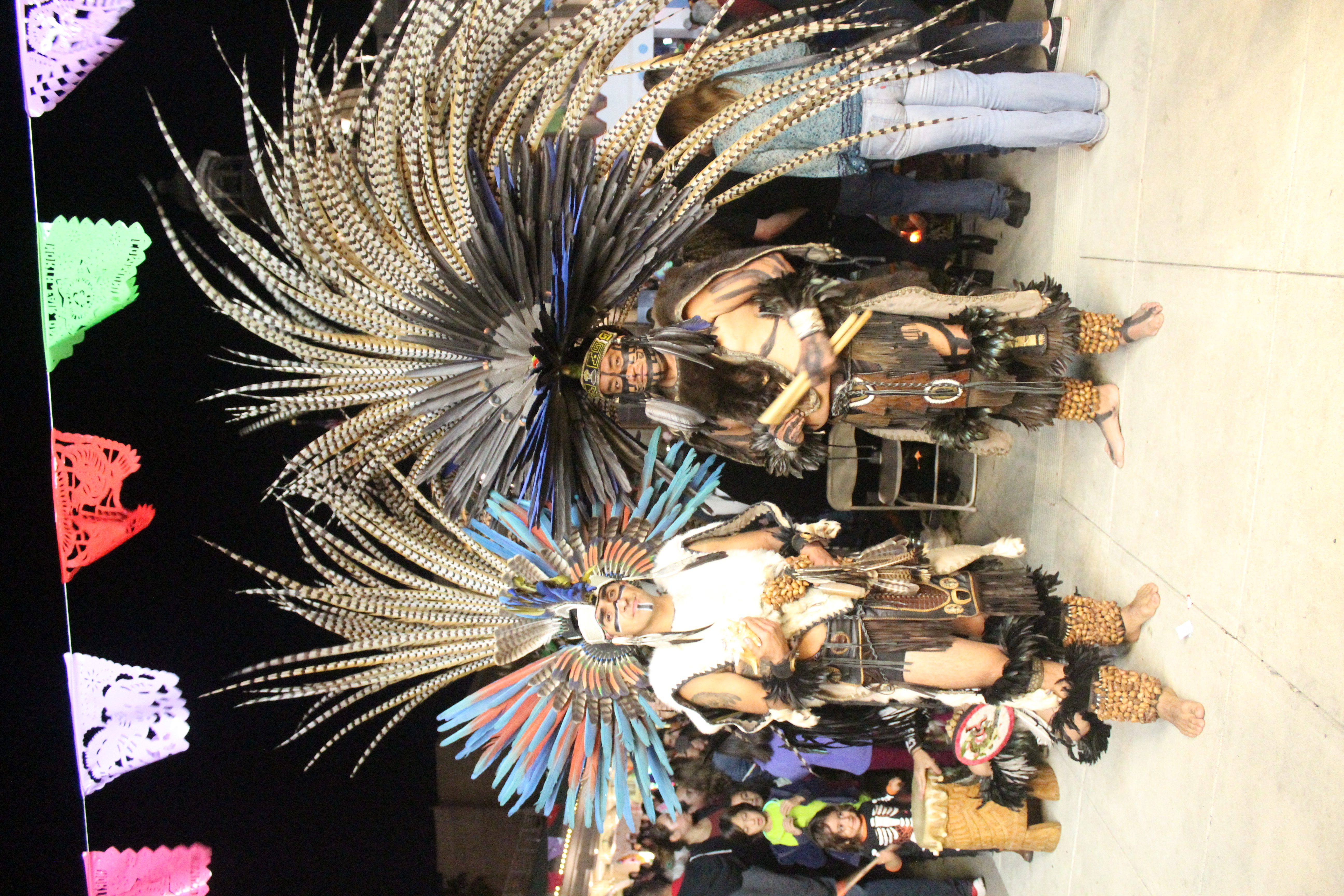 Aztec dancers with large feathered headdresses under papel picado at night